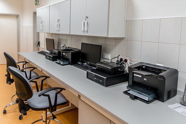kancelář, 2 židle u stolu, dva počítače, monitory a klávesnice a dvě tiskárny nad nimi bílé skříňky